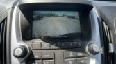 Car dashboard with a rearview camera display showing a parking lot with yellow guidance lines and the warning text 'Check Surroundings For Safety' at the bottom.