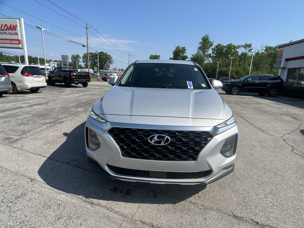 Front view of a silver Hyundai SUV in a dealership parking lot with other cars and signs in the background