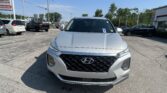 Front view of a silver Hyundai SUV in a dealership parking lot with other cars and signs in the background