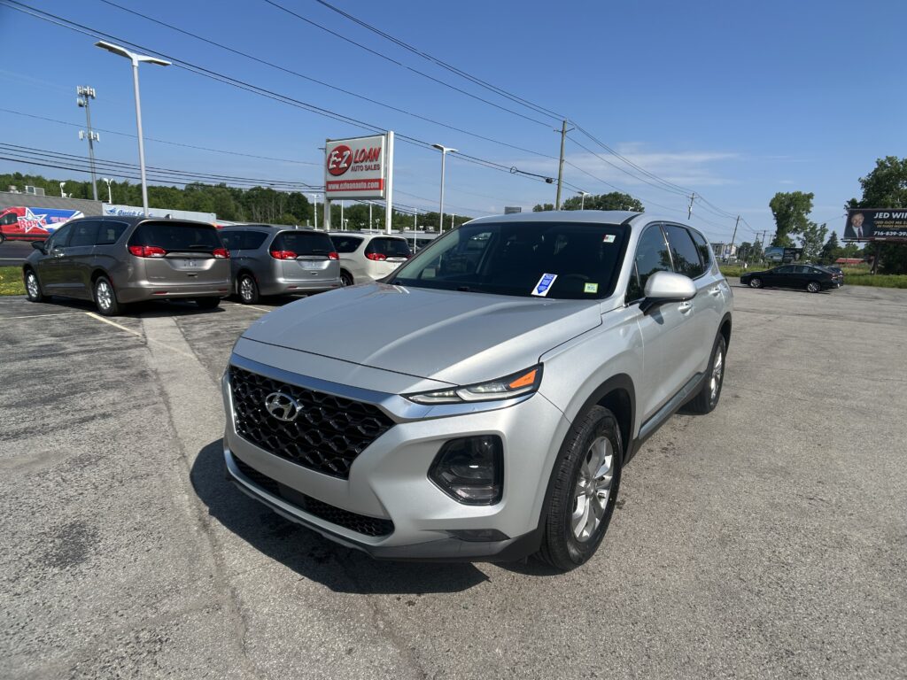 Front-angle shot of a silver SUV in a dealership lot with an EZ Loan Auto Sales sign and clear blue sky in the background (power lines visible).