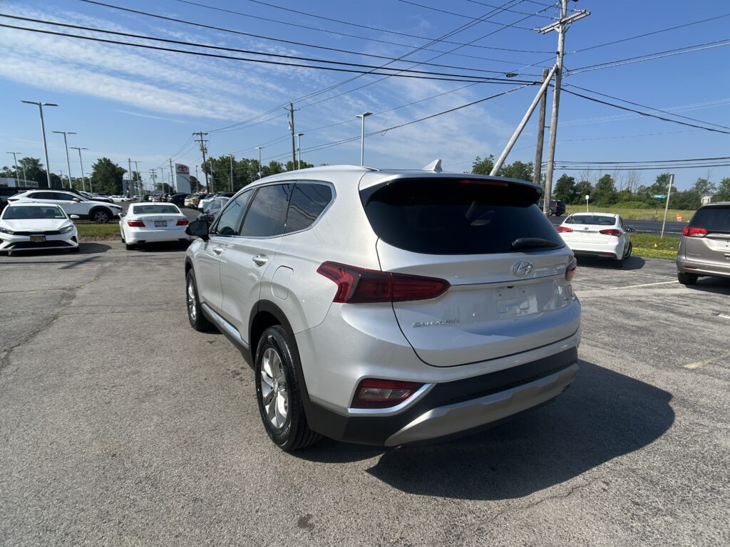 Rear three-quarter view of a silver Hyundai SUV in a busy dealership lot under a clear blue sky with power lines overhead when photographed from the back-right side of the vehicle safety view in the foreground.