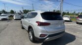 Rear three-quarter view of a silver Hyundai SUV in a busy dealership lot under a clear blue sky with power lines overhead when photographed from the back-right side of the vehicle safety view in the foreground.