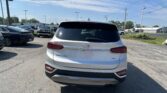 Rear view of a white Hyundai Santa Fe SUV in a crowded parking lot, with other cars and overhead power lines against a blue sky.