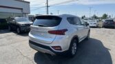 Rear view of a white Hyundai SUV in a car dealership lot with other cars in the background and a building on the left side of the frame.