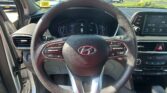 Driver's perspective inside a car: steering wheel and digital instrument cluster with an in-dash display visible, outside a parking lot of cars through the windshield.
