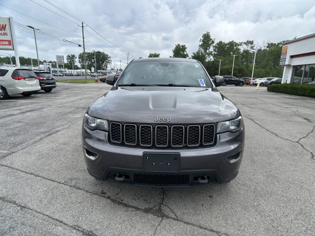 Front view of a gray Jeep SUV parked head-on in a busy dealership lot with other cars and buildings in the background.