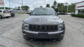 Front view of a gray Jeep SUV parked head-on in a busy dealership lot with other cars and buildings in the background.
