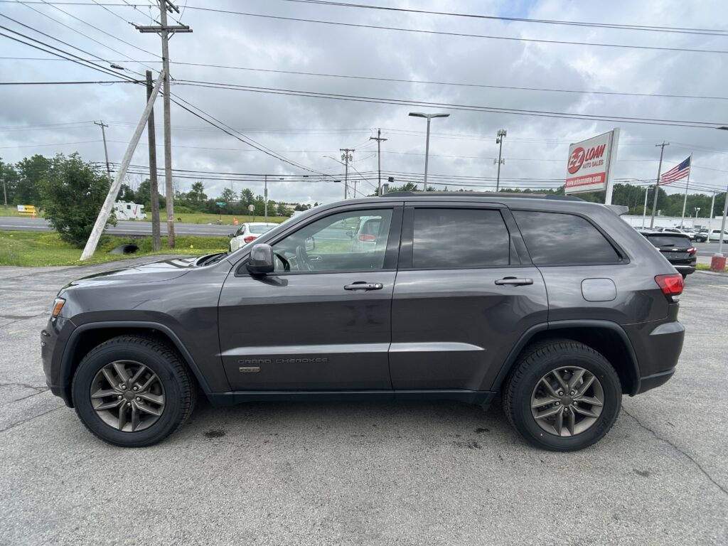 Gray Jeep Grand Cherokee in a parking lot beside a roadside auto sales lot with an EZ LOAN sign and American flag in the background.