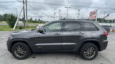 Gray Jeep Grand Cherokee in a parking lot beside a roadside auto sales lot with an EZ LOAN sign and American flag in the background.