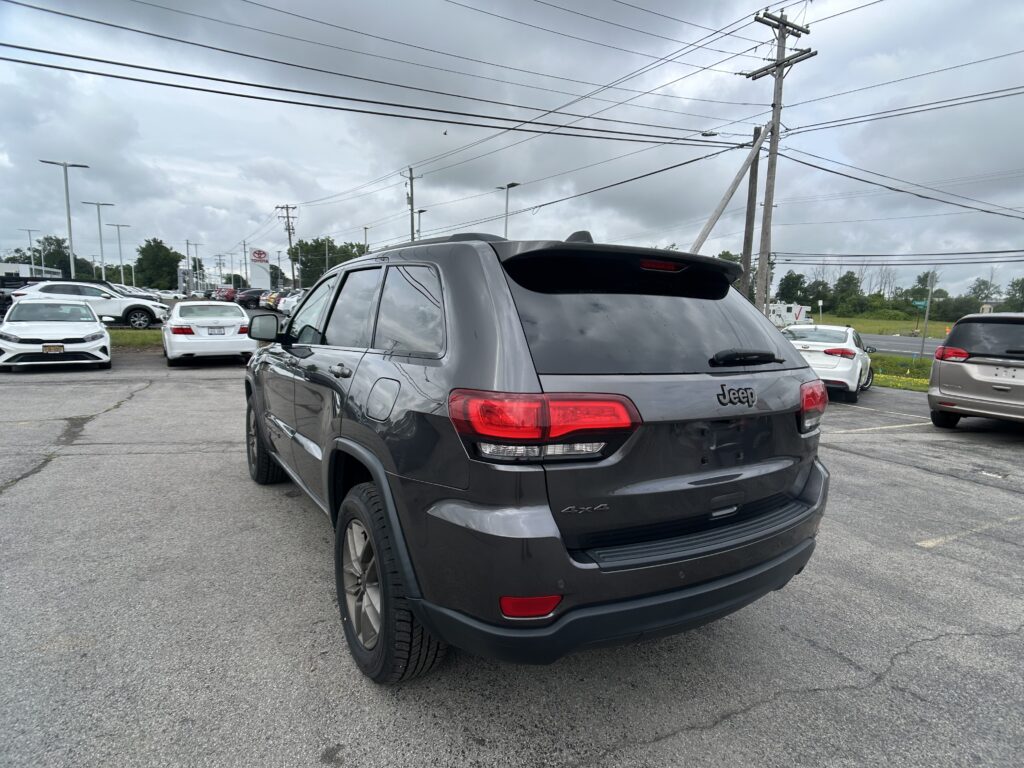 Rear view of a gray Jeep SUV in a busy car dealership lot on a cloudy day, with other cars parked nearby and overhead power lines.