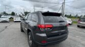 Rear view of a gray Jeep SUV in a busy car dealership lot on a cloudy day, with other cars parked nearby and overhead power lines.