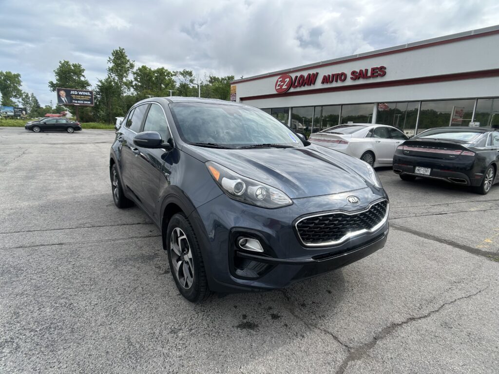Gray Kia SUV parked in foreground outside EZ Loan Auto Sales dealership with other cars and a billboard in the background.