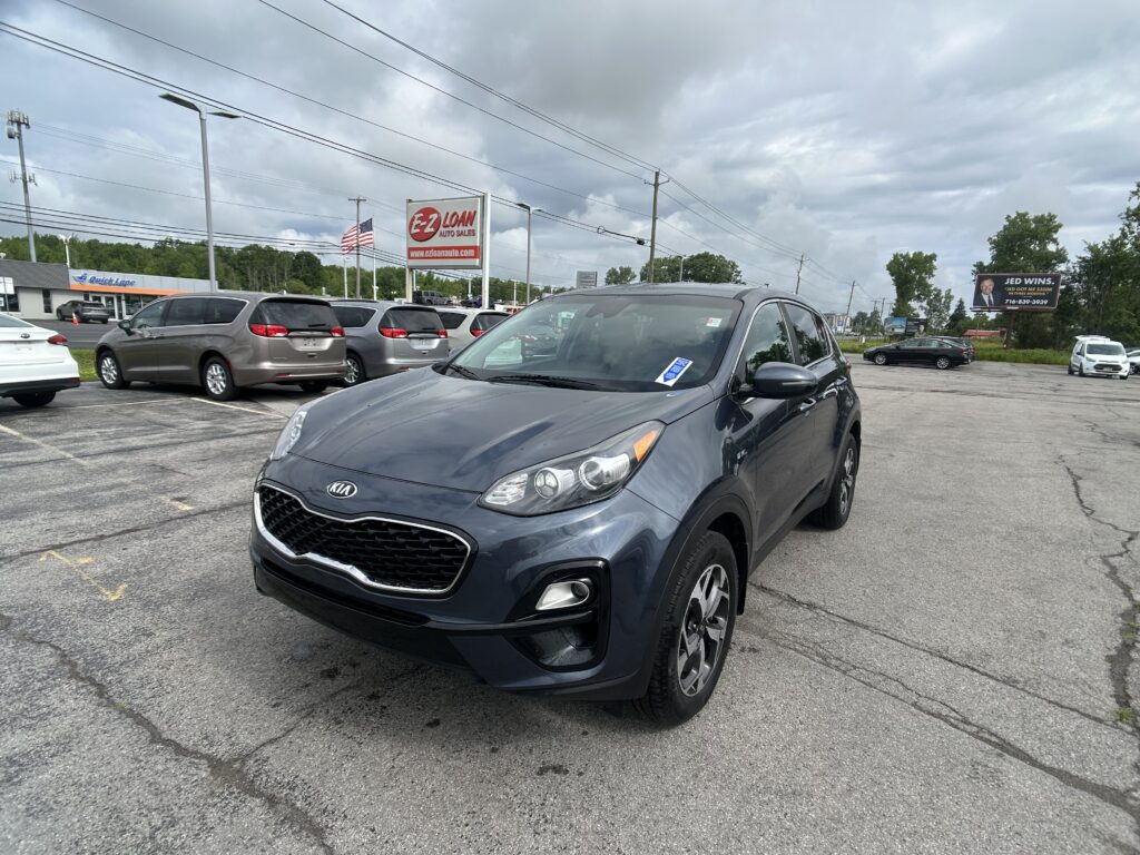 Gray Kia SUV in the foreground at a car dealership lot with other minivans parked nearby and an EZ Loan Auto Sales sign in the background, under a cloudy sky.