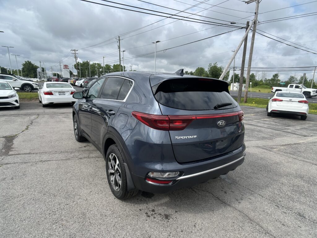 Blue Kia Sportage in a dealership parking lot, seen from behind, with other cars and overhead power lines against a cloudy sky.