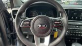 Driver’s-seat view of a Kia steering wheel and dashboard, with an in-car infotainment screen and exterior cars visible through the windshield.