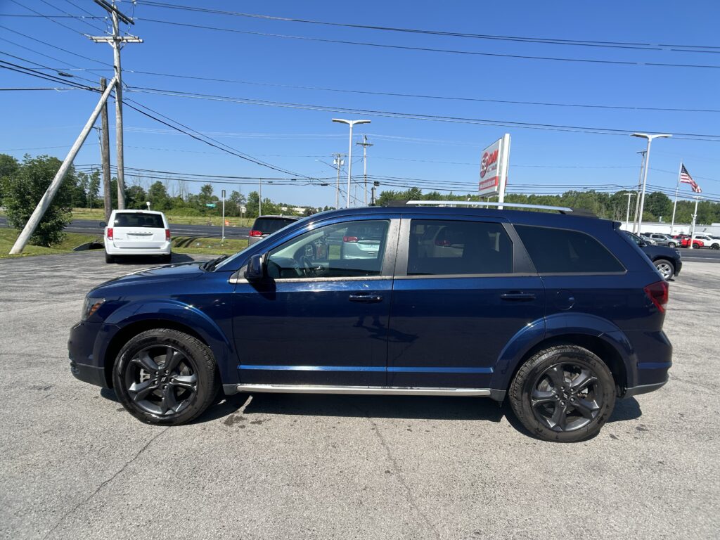 Blue SUV in a dealership lot with other cars, power lines overhead, and a distant American flag on a clear day.