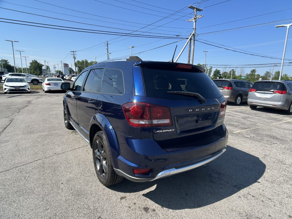 Blue Dodge Journey SUV parked in a busy dealership lot with other cars and overhead power lines under a blue sky.