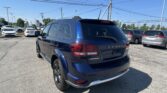 Blue Dodge Journey SUV parked in a busy dealership lot with other cars and overhead power lines under a blue sky.