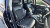 Inside view of a dark blue SUV with black leather seats, front and rear seats visible, center console with cup holders, and the driver's door open; outdoors you can see a dealership lot through the windows.