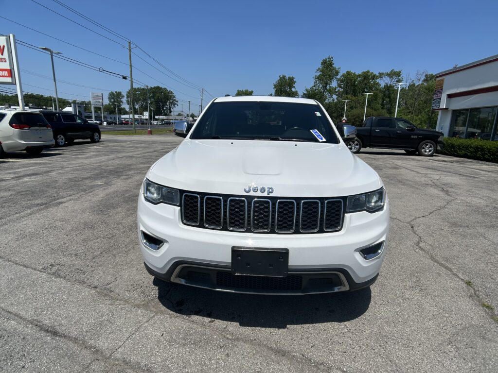 Front view of a white Jeep SUV in a car dealership lot with other vehicles and a building in the background.