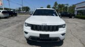 Front view of a white Jeep SUV in a car dealership lot with other vehicles and a building in the background.
