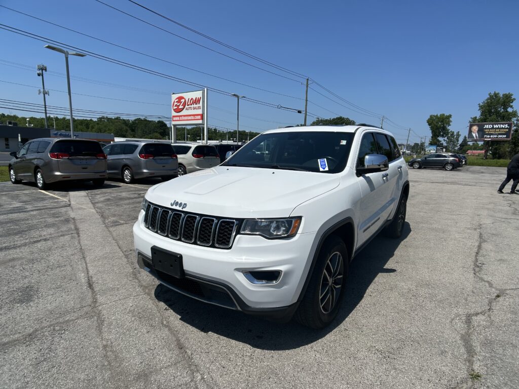 White Jeep SUV in a car dealership lot with other vans parked and an EZ LOAN Auto Sales sign in the background on a clear blue sky day, front-right view.