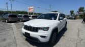 White Jeep SUV in a car dealership lot with other vans parked and an EZ LOAN Auto Sales sign in the background on a clear blue sky day, front-right view.