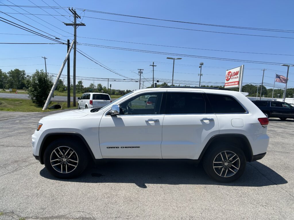 White Jeep Grand Cherokee parked in a dealership lot with an EZ LOAN Auto Sales sign and American flag in the background.