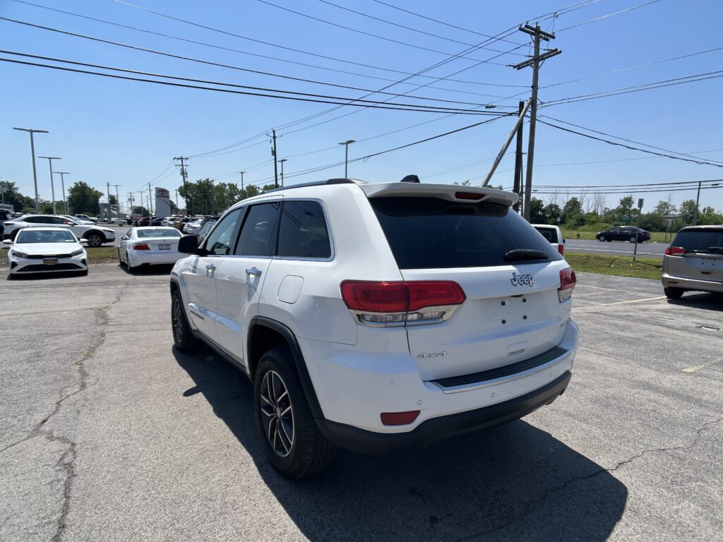 White Jeep Grand Cherokee SUV in a dealership parking lot with other cars and tall power lines overhead against a clear blue sky