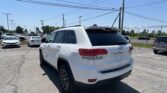 White Jeep Grand Cherokee SUV in a dealership parking lot with other cars and tall power lines overhead against a clear blue sky