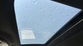 Interior of a car with a large sunroof open to a clear blue sky, raindrops visible on the glass.