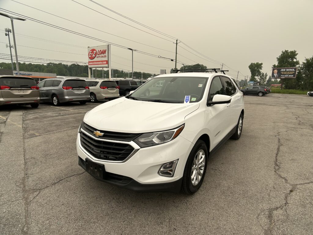 White Chevrolet SUV in the foreground at a dealership lot with several minivans parked behind and an EZ Loan sign in the distance.
