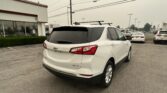 White Chevrolet SUV with roof rack seen from the rear in a dealership parking lot, other cars and a Toyota dealership sign in the background