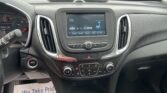 View from the driver's seat of a car's interior focusing on the center touchscreen/console with parked cars visible through the windshield outside. The screen shows AM radio and a blue tower icon, and the gear shifter is in the foreground.