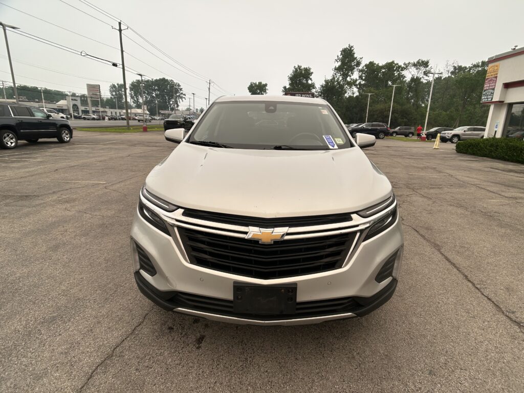 Front view of a white Chevrolet SUV in a car dealership lot with other vehicles and a building in the background]","Front close-up of the white Chevrolet SUV at a dealership, with signs and cars in the lot behind
