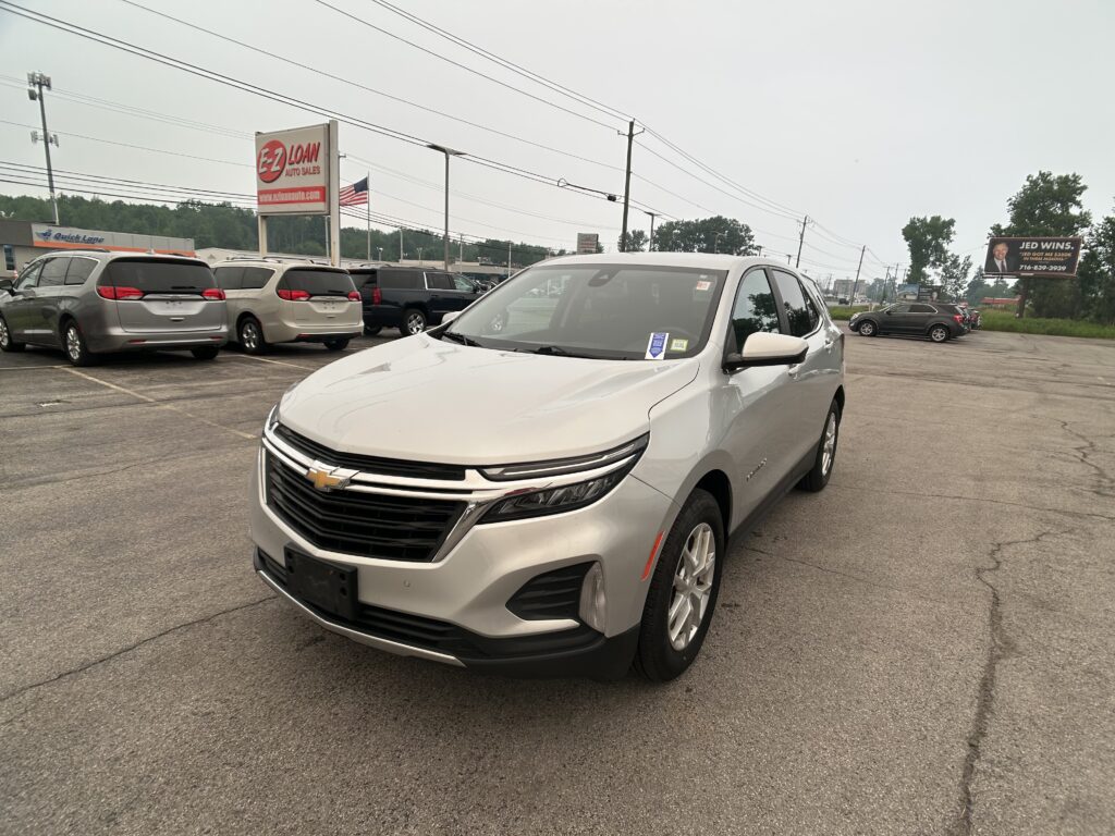 White Chevy in the foreground at a car dealership lot with other vans and an EZ LOAN Auto Sales sign nearby, plus a billboard in the distance.