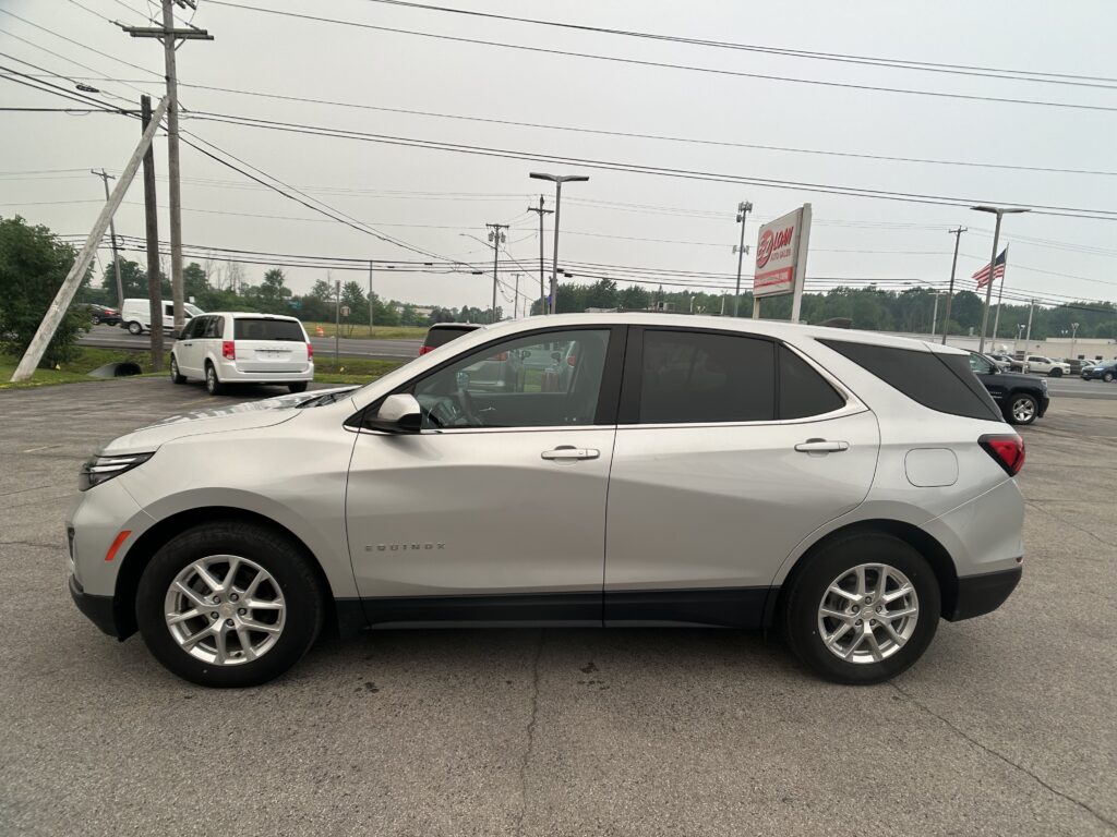 Silver Chevrolet Equinox parked in a dealership lot with power lines overhead and an American flag in the background.
