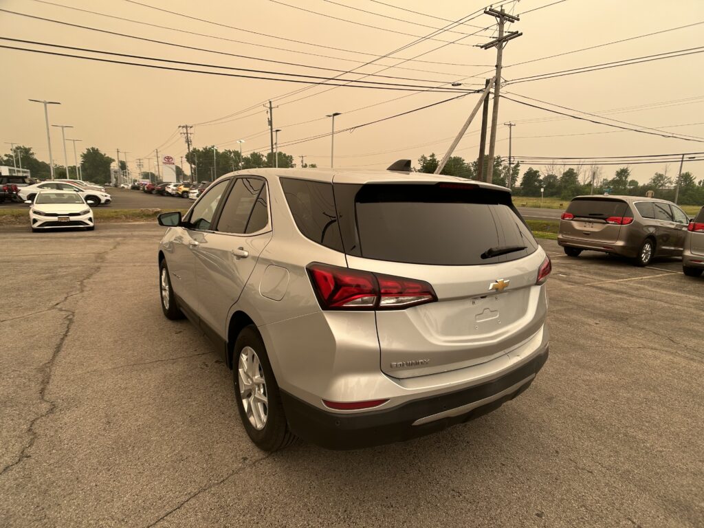 Silver Chevrolet SUV (Equinox) in a parking lot with multiple cars and power lines overhead, dealership in the background.