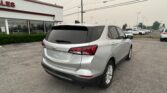Rear view of a silver Chevrolet SUV parked in a car dealership lot, with a glass-front showroom and a row of cars in the background.