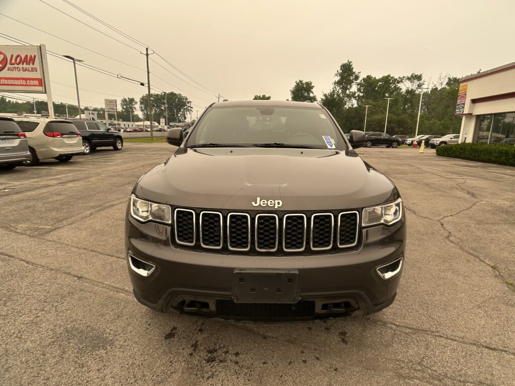 Front view of a dark gray Jeep SUV parked in a dealership lot, with other cars and a sign visible on the left.