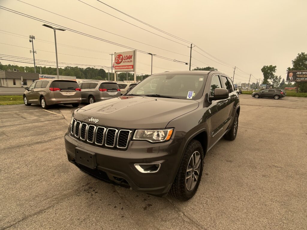 Front three-quarter view of a gray Jeep SUV parked in a lot with other minivans and a large E-Z LOAN Auto Sales sign in the background under a pale sky.