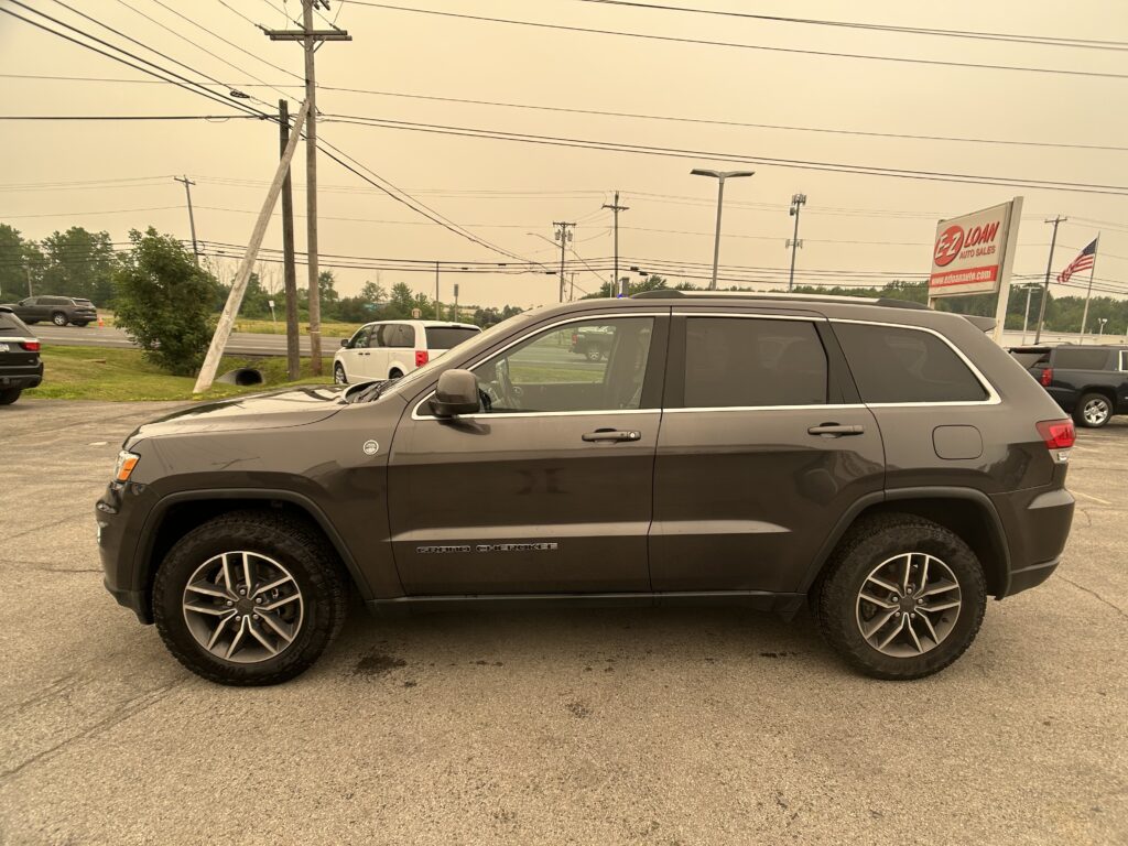 Side view of a gray Jeep Grand Cherokee in a parking lot with power lines and a roadside EZ Loan Auto Sales sign in the background.