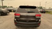 Rear of a gray Jeep SUV in a car dealership parking lot with other cars and power lines overhead on a hazy day.