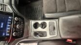 Interior of a car cabin focused on the center console: gear shifter, cup holders, climate controls, and a nearby passenger seat with a fabric upholstery. A floor mat and a paper lie on the floor.