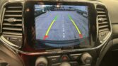 Car infotainment screen showing rearview camera with yellow and red guiding lines and 'Check Entire Surroundings' message on a parking lot backdrop.