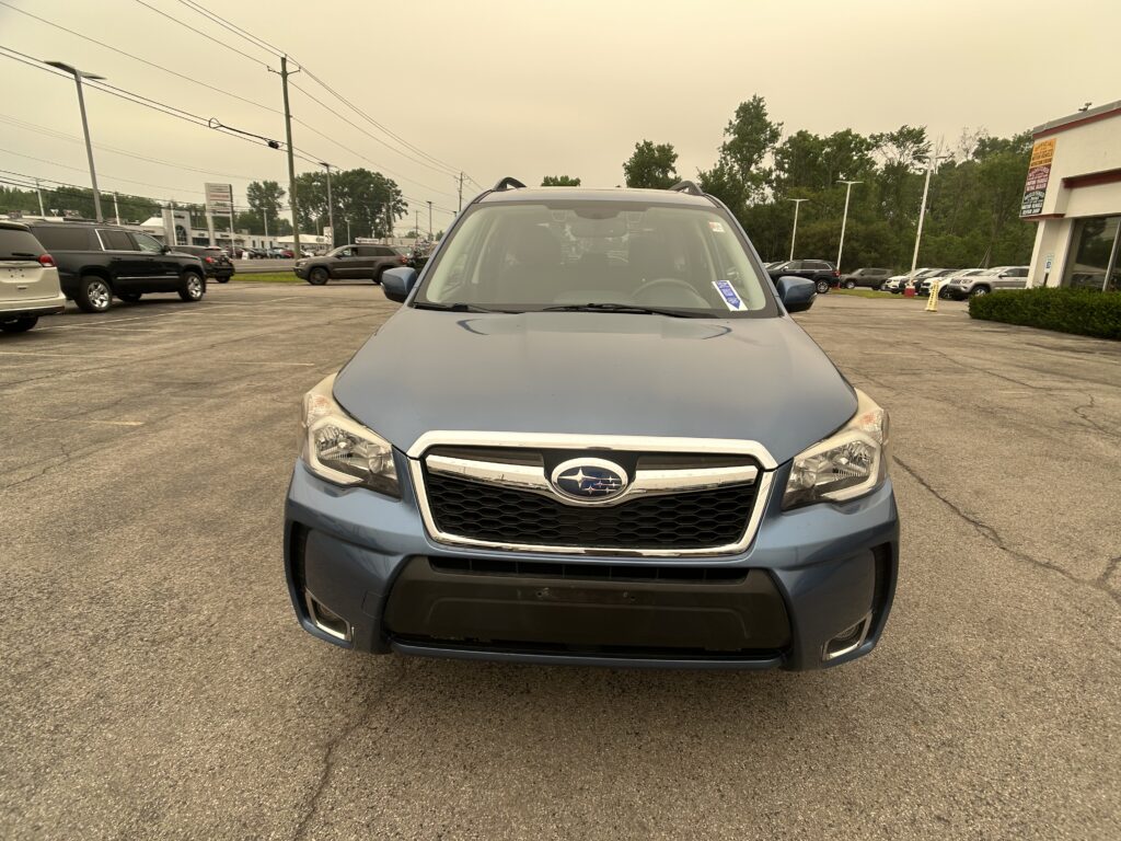 Front view of a blue Subaru SUV in a parking lot, dealership buildings and other cars in the background under a pale sky.