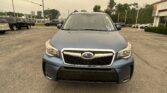 Front view of a blue Subaru SUV in a parking lot, dealership buildings and other cars in the background under a pale sky.