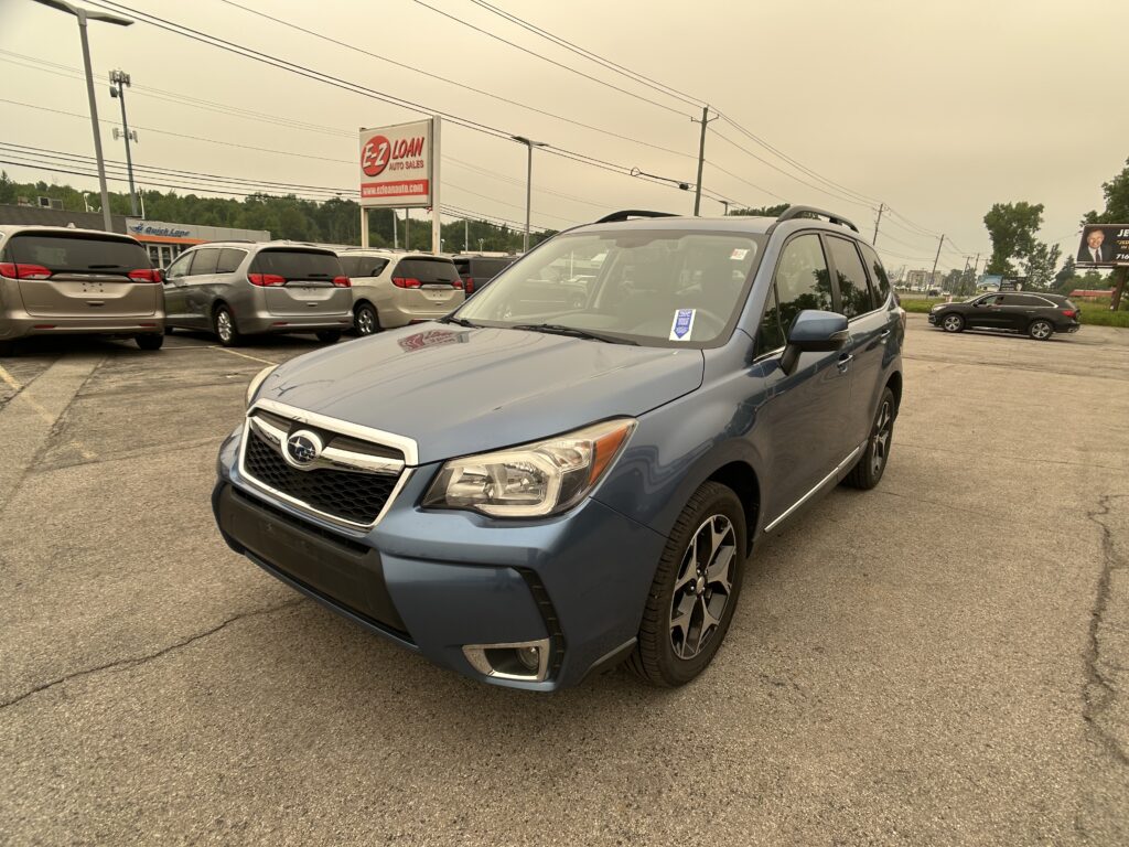 Blue SUV in the foreground at a car dealership lot with several minivans parked behind and power lines overhead under a cloudy sky, sign reading 'E-2 LOAN Auto Sales' nearby.