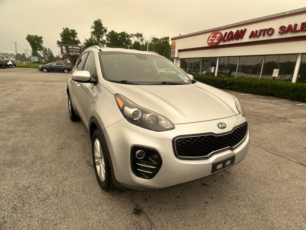 Silver Kia SUV parked in a lot in front of an EZ Loan Auto Sales building with large windows and red sign.