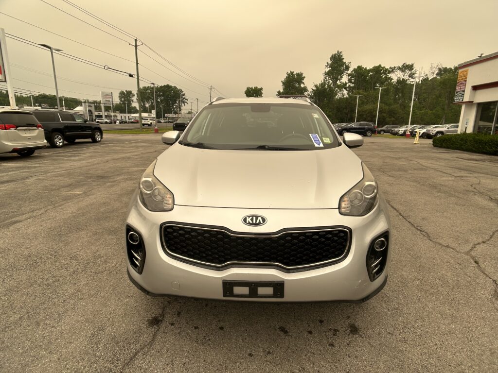 Front view of a silver Kia SUV parked in a dealership lot with other vehicles and a building on the right side.